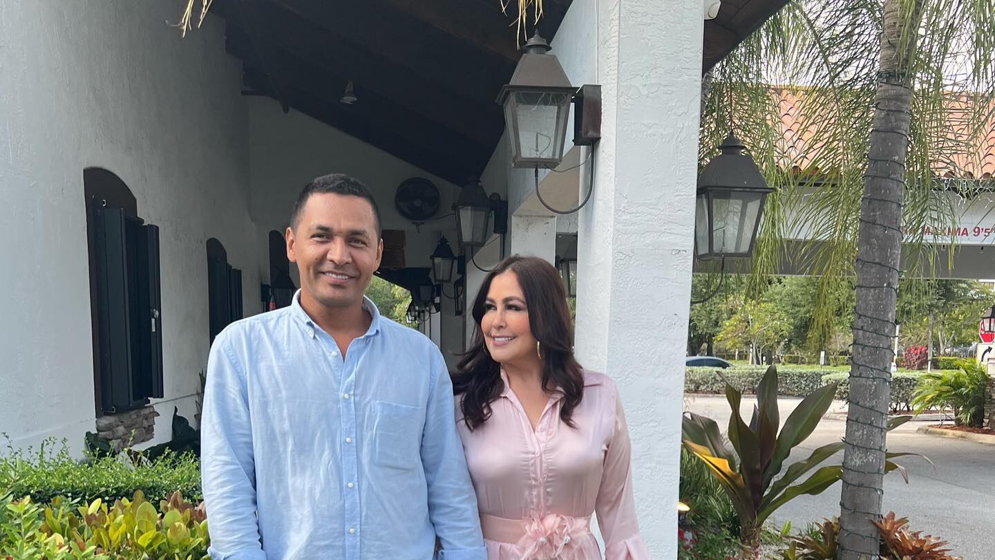 Tomados de la mano Arelys Henao y su esposo con una sonrisa plena en el rostro de ambos en medio de uno de los corredores de una casa de campo