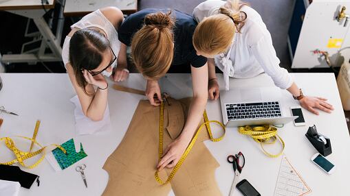 Tres mujeres diseñadoras de moda trabajando con un patrón de costura. Vista desde arriba con una cinta métrica, una laptop y un teléfono celular sobre la mesa. (Referencial)